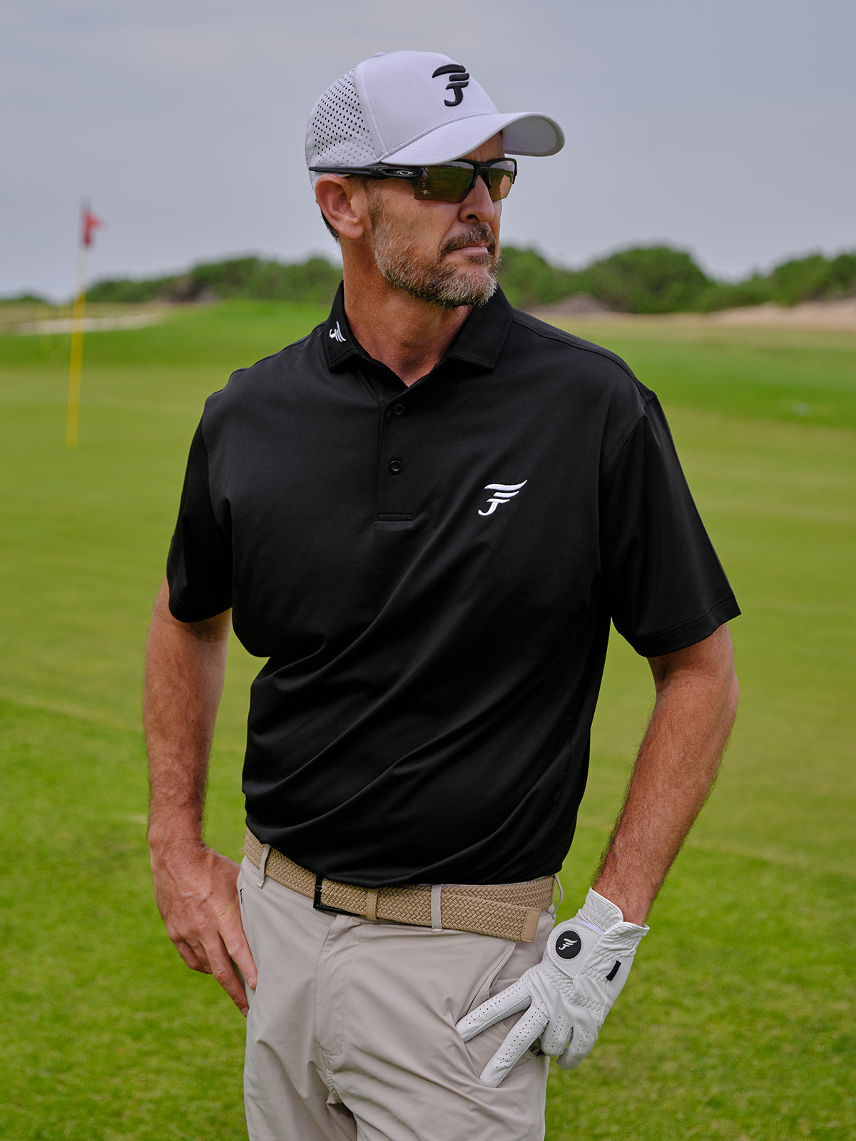 Man on a golf course wearing a black shirt, beige pants, and a white cap with a number 3.