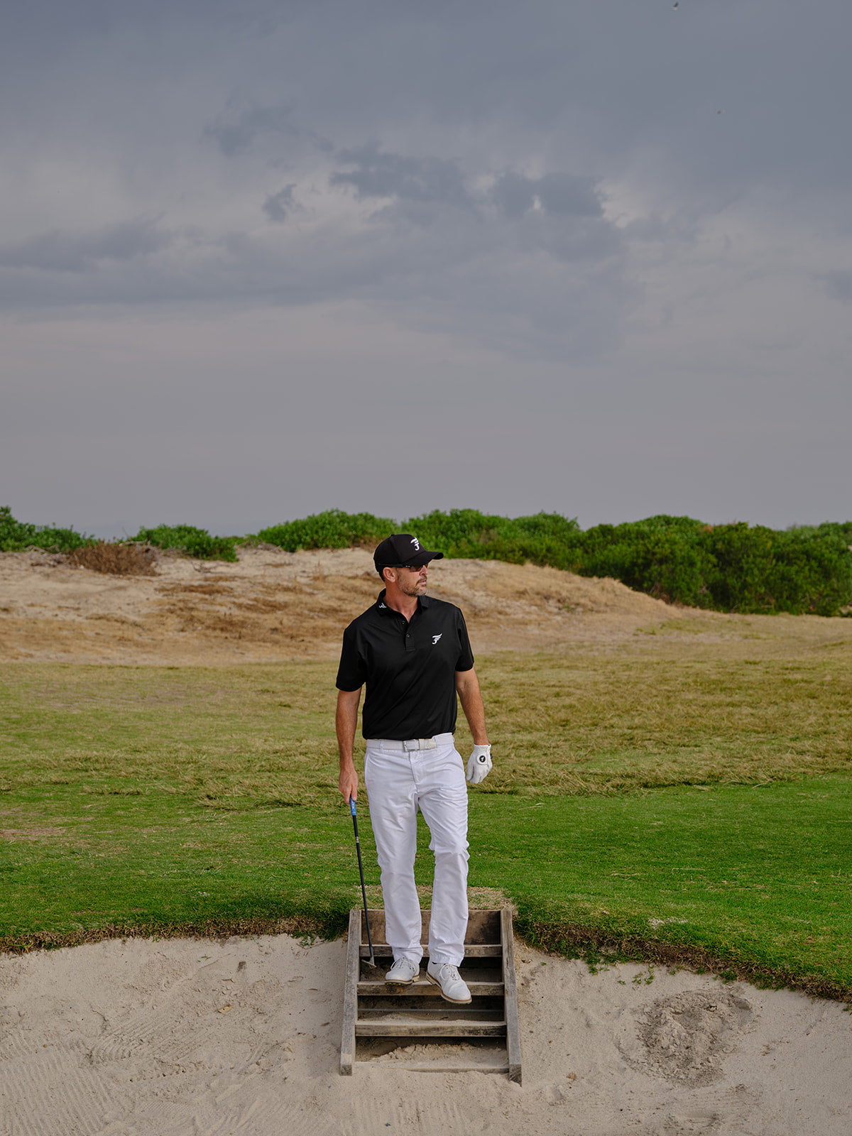 Golfer standing on a sand trap with a golf club, wearing a black shirt and white pants.
