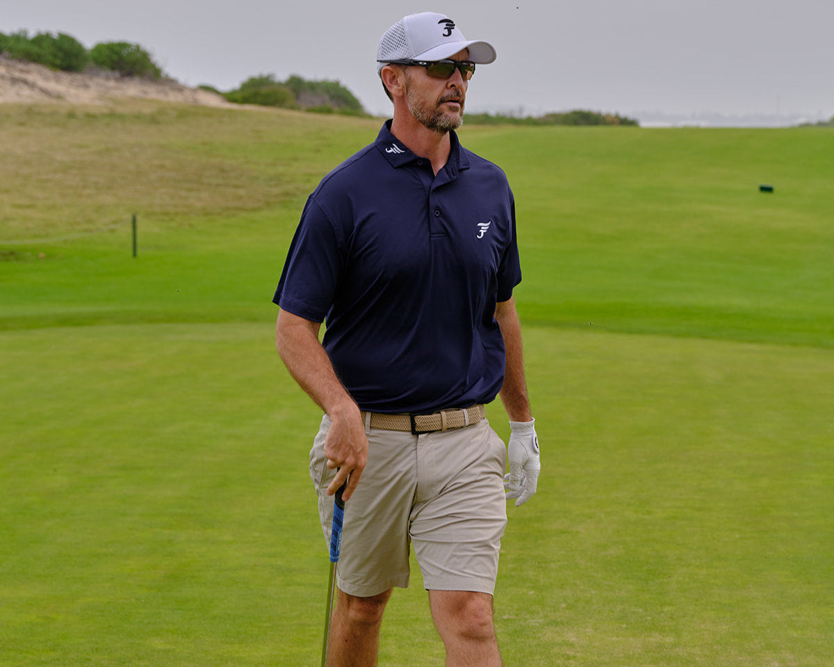 Man in navy blue polo shirt and beige shorts standing on a golf course holding a club.